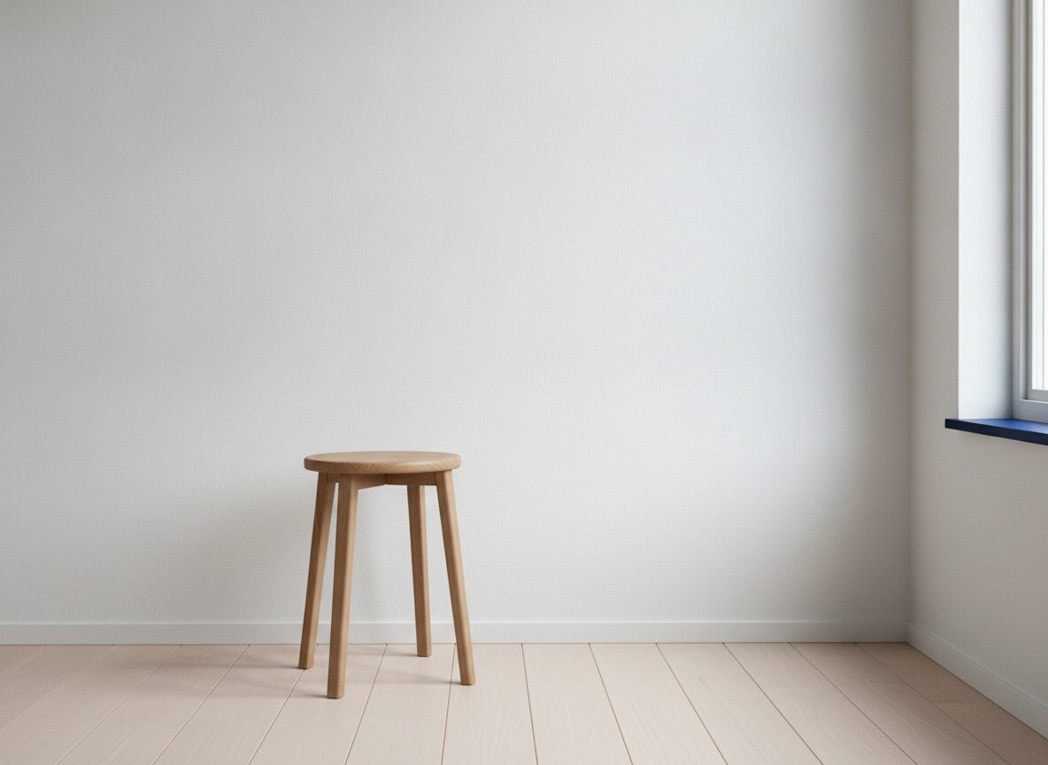 A round-seat oak stool with four straight legs stands on a pale wood plank floor in front of a plain white wall, with a window and a dark blue sill at the right edge letting in soft daylight.
